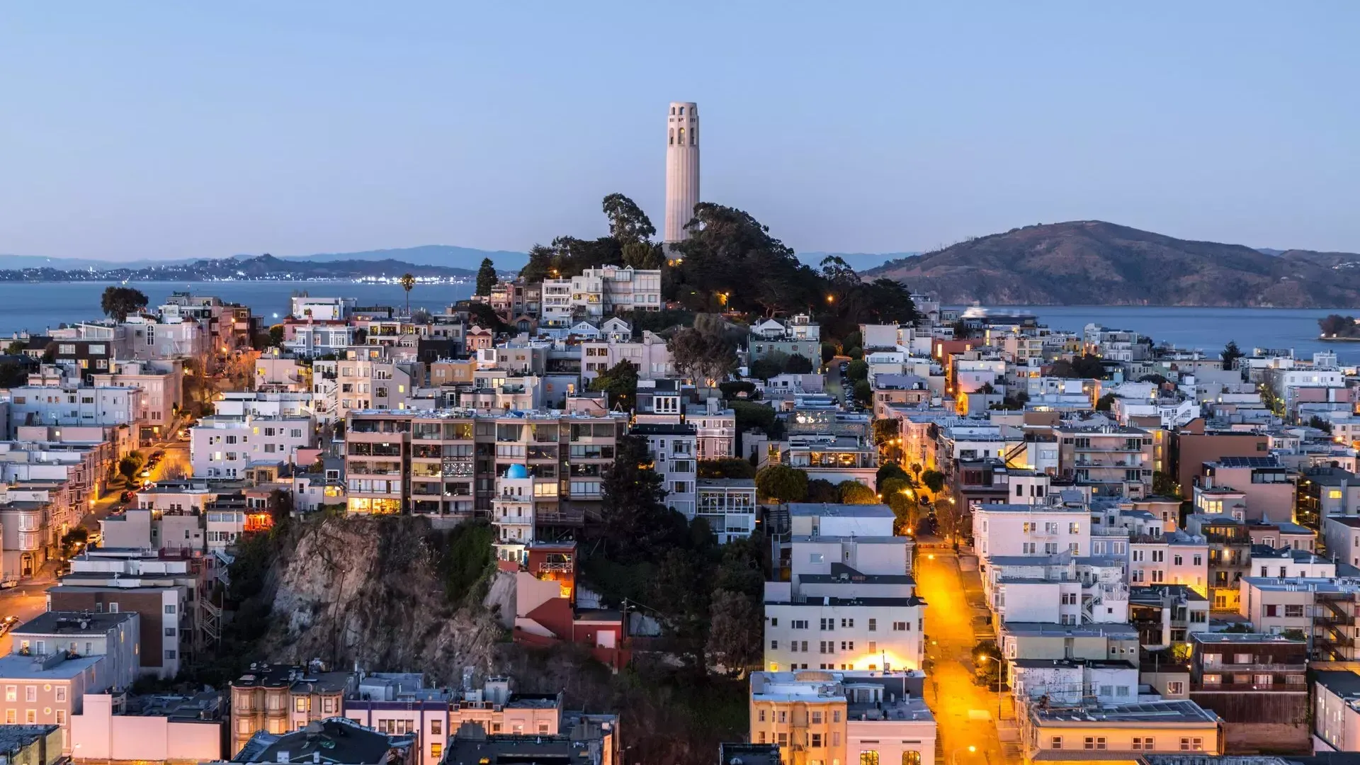 San Francisco's Coit Tower at dusk, with lighted streets before it and the San Francisco Bay behind it.